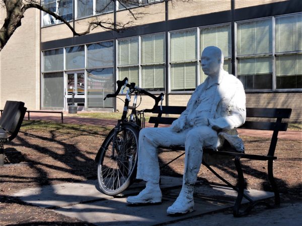 A tour bike leaning on a bench on which an all white sculpture of an African American man sits.