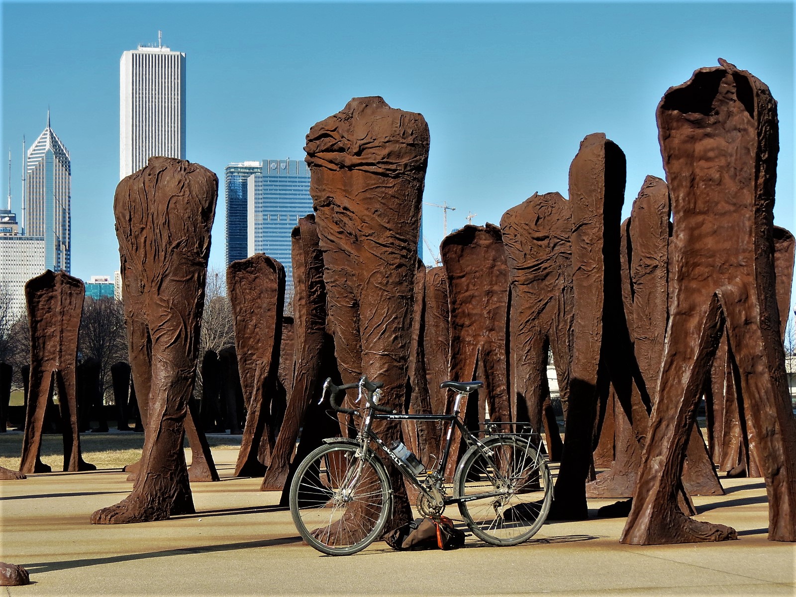 A tour bike leaning on a brown metal sculpture of many armless human like striding figures with skyscrapers in the background