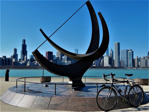 A tour bike leaning in front of a sun dial sculpture with the Chicago skyline in the background.