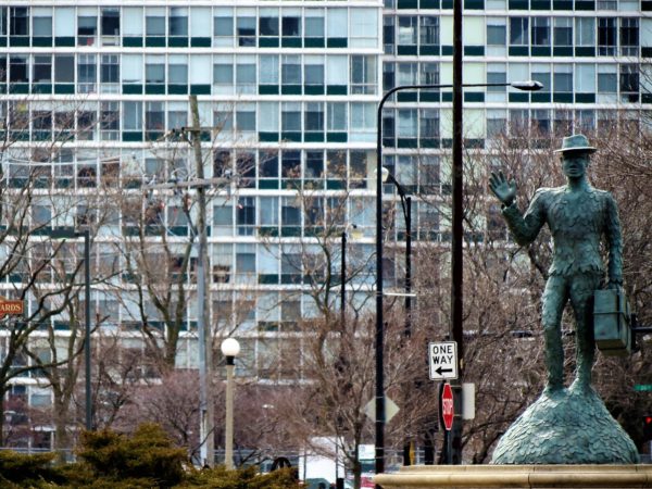 A bronze sculpture of an African American man in a hat and shoe sole coat with a bag in one hand while the other is waving, the glass and steel of Prairie Shores stands as a wall behind.