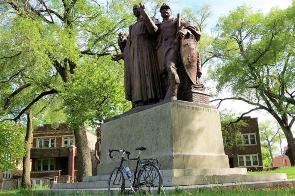 A tour bike leaning in front of a bronze sculpture of a Native American, a priest, and a soldier with leafing trees behind