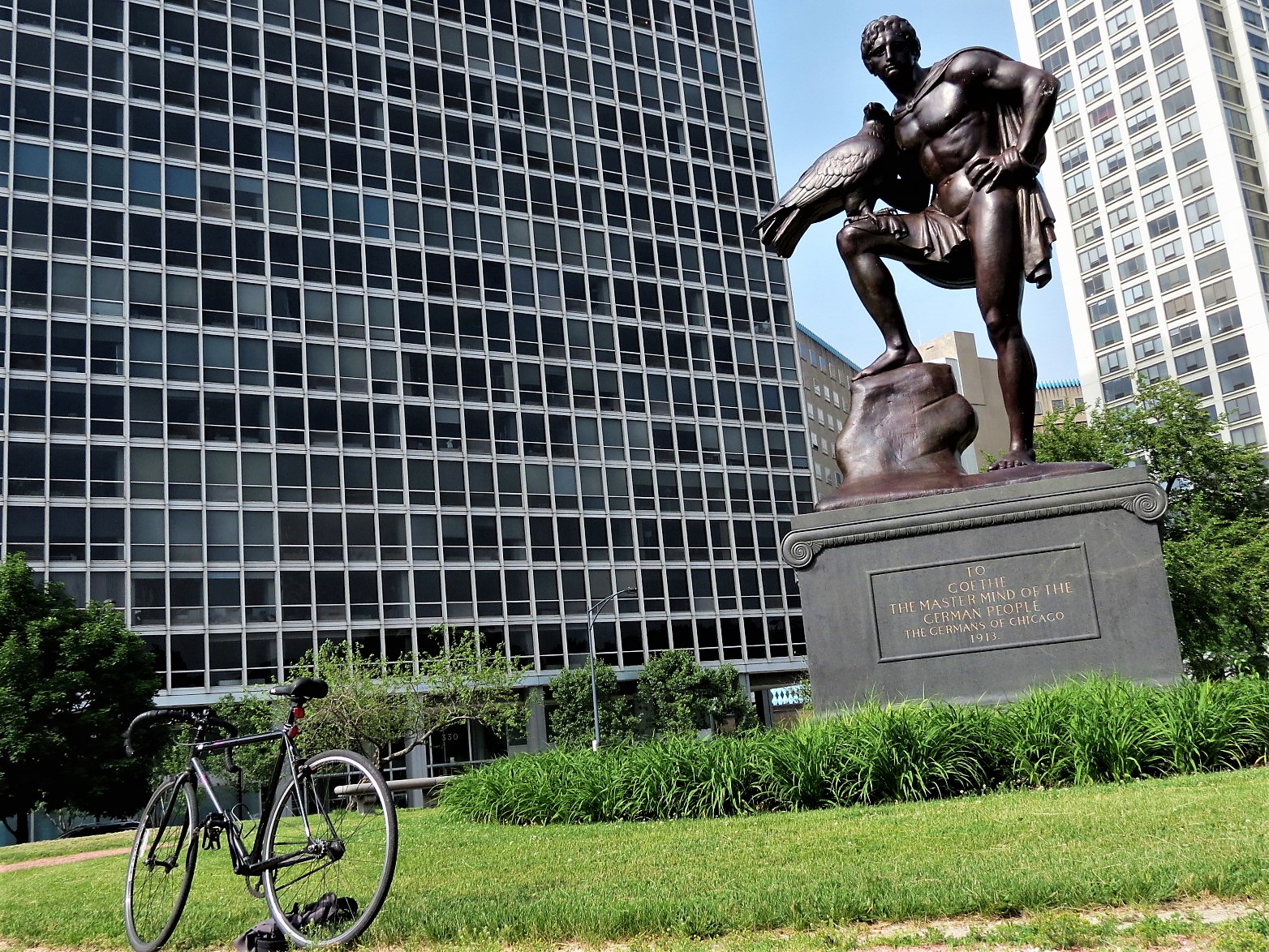 A tour bike standing in front of a bronze sculpture of a figure given the stature of a Greek god holding a bird in one hand and a foot propped on a rock with the glass facade of a condo building behind.