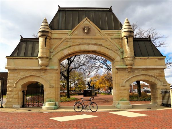 A tour bike standing in front of a yellow limestone neo-Gothic 1860s gate structure with a cow's head.