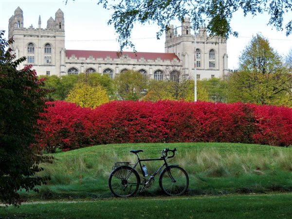 A tour bike standing in front of a small green hill with low red leaf trees behind with pale green trees behind with Neo-Gothic limestone building behind.