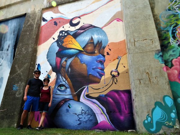 Two CBA bike tour riders posing in front of a mural of a dark skinned woman with blue pain across her face, butterfly wings in her hair, an eye in her neck, and a hanging key
