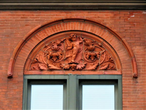 Thin red brick arch on a red brick wall with a floral and cupid red terra cotta detail.