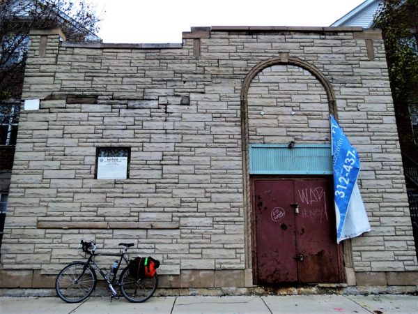 A tour bike leaning fake gray stone siding with a high stone arch with a red rusted locked metal door and banner hanging by one corner.