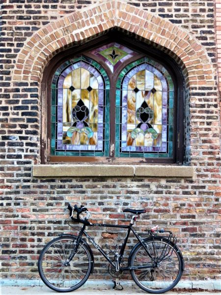A tour bike leaning on a Chicago common brick wall with a window of two pains of stained glass window from the inside.