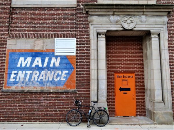 A tour bike leaning on a red brick building with an art deco stone doorway with two Main Entrance signs pointing in opposite directions.