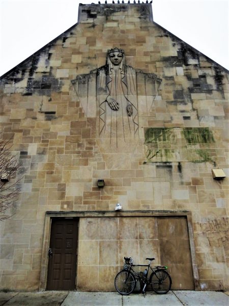 A tour bike leaning on the back side of a building with black and green stained stone and a Virgin Mary bas relief.