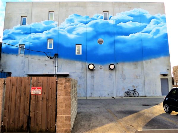 A tour bike leaning on a gray back wall with a large blue painted cloud towards the top.