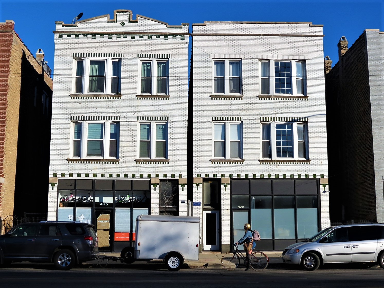 A Chicago bicyclist riding past two three story white and green glazed brick buildings.