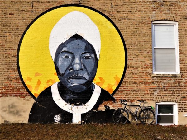 A tour bike leaning on a mural of a black woman in a white head wrap and white piped black dress with a bright yellow sun or halo behind.