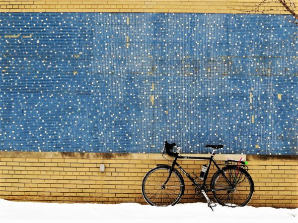 A tour bike in the snow leaning on a yellow brick wall with large band of blue and white small square tiles.