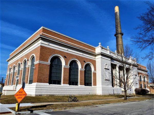 A tour bike on the sidewak in front of the large block tan brick and white stone Central Avenue Pumping Station with large arched windows and smokestack.