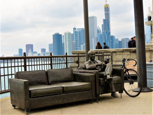A tour bike leaning on a sculpture of a man sitting on a couch with skyscrapers in the background.