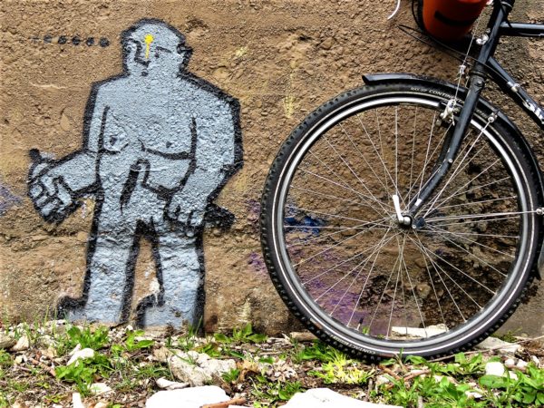 A bicycle wheel next to aa mural of a little gray man with a spray can.