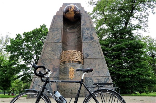 A tour bike in front of a dark marble pyramid shape with gold metal Art Deco text and a black and gold globe