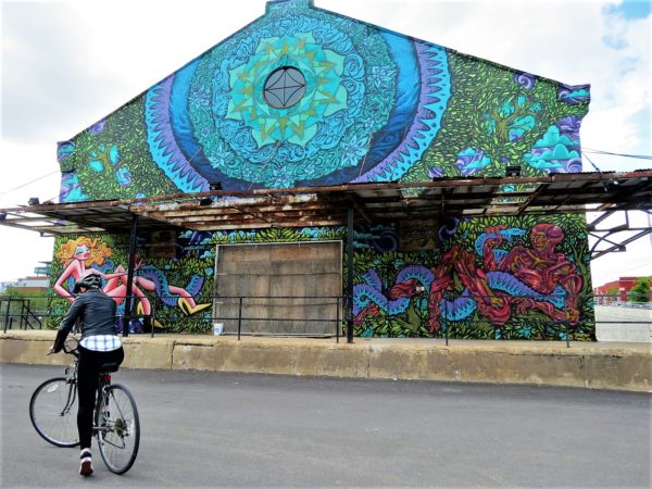 A CBA bike tour rider in front of a Fulton Market warehouse with a large blue, purple, and green Adam and Eve in the Garden of Eden mural by Solomon Souza and Jas Petersen.