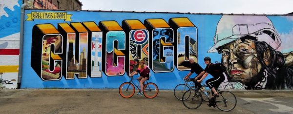 Three CBA bike tour riders cycling in front of the Logan Square Greetings from Chicago and female hard hat worker mural.
