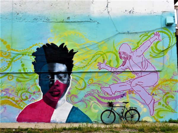 A tour bike leaning on a mural of green and pink ribbons, a dark outlined pink dancing figure and a stylized black young man