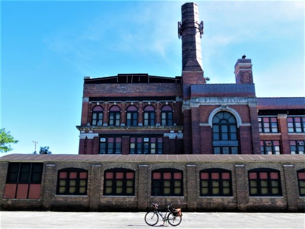 A tour bike standing in front of a red and tan brick early 1900s school with many windows and a smokestack.