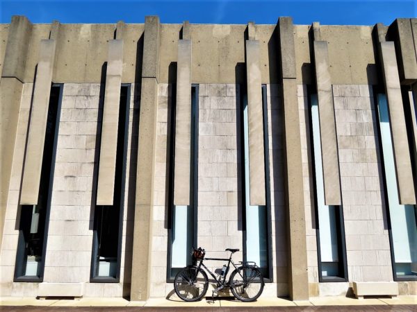 A tour bike leaning on a Brutalist style gray concrete wall with vertical segments covering long thin vertical windows.