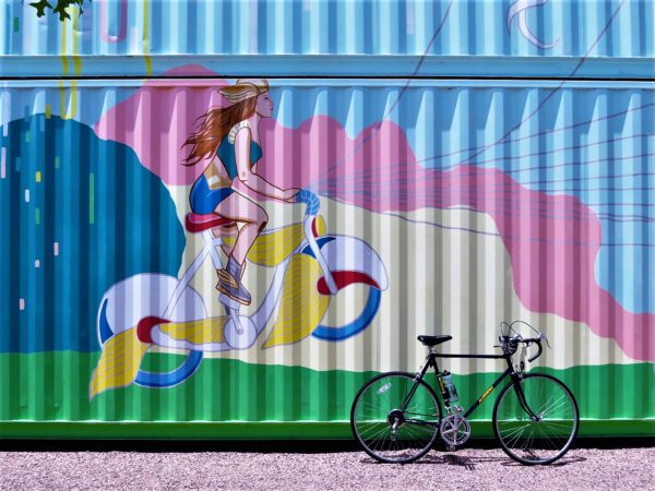 A tour bike leaning on a pastel color shipping container mural of a woman with a winged helmet and boots riding a winged bicycle.