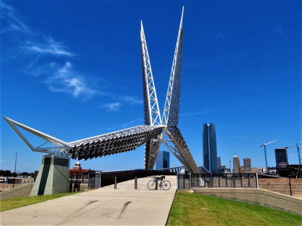 A tour bike standing below a metal scissor tail fly catcher sculpture with the OKC skyline in the background.