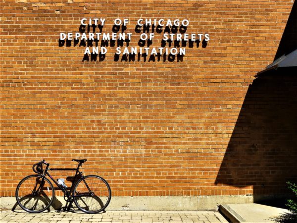 A tour bike leanning on a tan yellow brick wall with a metal type sign accented by the heavy shadow of the letters.