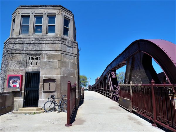 A tour bike leaning on the grey stone art deco minder house of a maroon painted pony truss bridge.