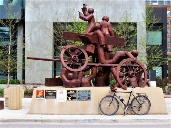 A tour bike leaning in front of a red copper statue of faceless igures building a barricade of wheels and planks