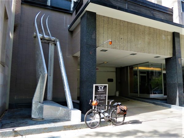 A tour bike standing in front of a metal swing Swiss Army knife like sculpture with three blades raised.