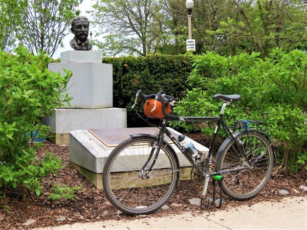 A tour bike leaning in front of a bronze male bust sitting on a white stone pedestal between green leafed bushes..