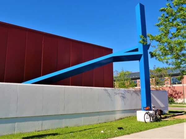 A tour bike leaning on the concrete base of a blue metal sculpture with two large bars, on vertical one diagonal connected by another bar.