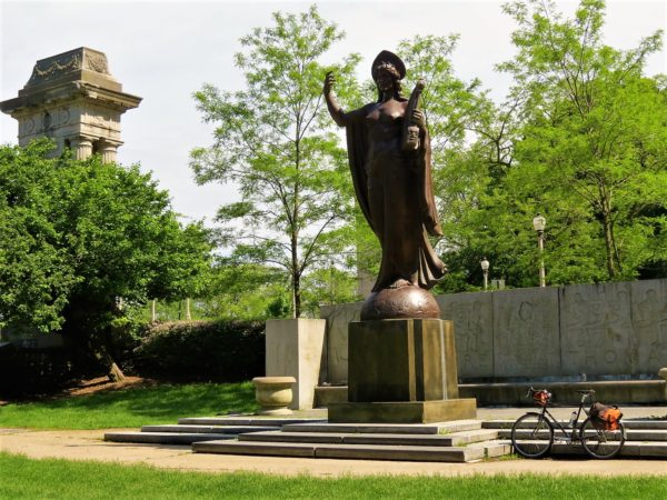 A tour bike leaning next to a bronze statue of a woman in robe and head covering holding a harp with green trees and a Neo-classical gate behind.