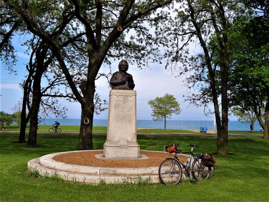 A tour bike leaning on the dias below the pedestal of a bronze man's bust with trees, lake, and bike rider in the background.