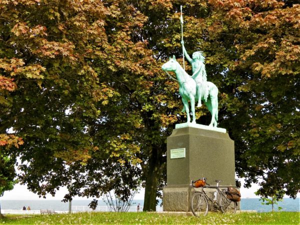 A tour bike leaning on the pedestal of a bronze sculpture of an American Indian in a headress on a horse holding a spear vertically with a background of brownish red leaves.