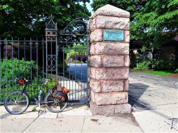 A tour bicycle leaning on the black metal fence next to a stone gate pillar with a bronze plaque that says Graceland Cemetery.