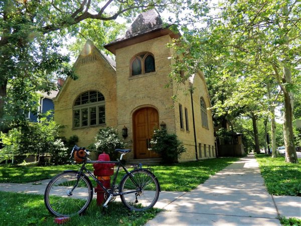 A tour bicycle leaning in front of a small yellow brick former church converted home with a two story tower and framed by green trees.