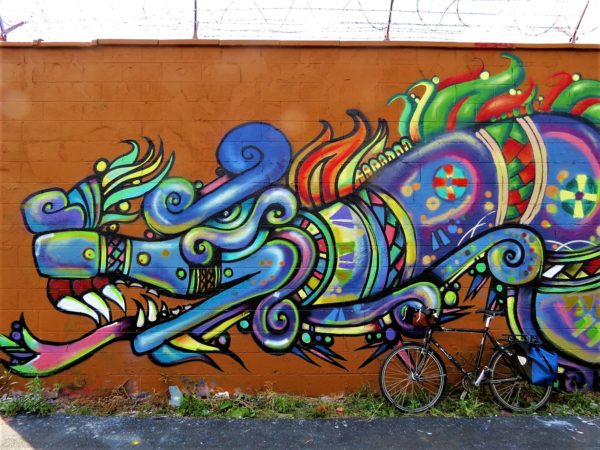 A tour bicycle leaning on a mural of a multi-colored Aztec style dragon like creature on an ochre background.