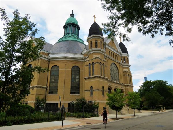 A CBA tour rider riding passed a yellow brick and patina copper dome church.