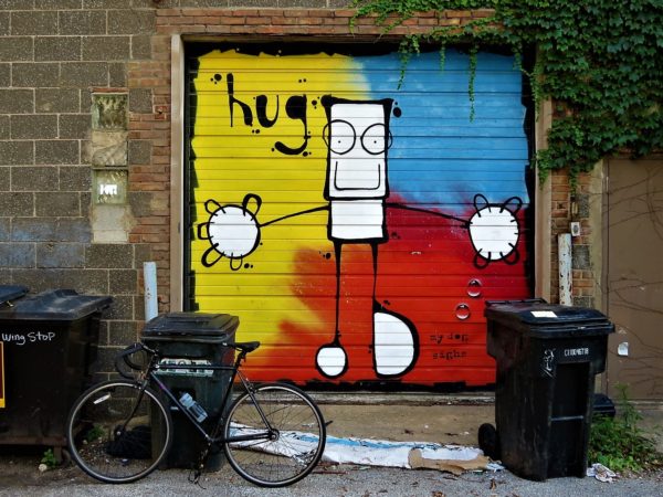 A tour bike leaning on a black plastic trash can in fornt of a roll up garage door painted with a mural of a rectangular figure with arms wide on a tripartite blue, yellow, a red background.