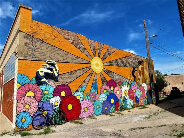 A tour bike leaning on a building back wall mural of multicolored flowers, a stature covering its face, and a sun with thick orange rays.
