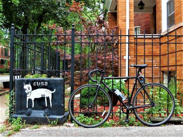 A tour bicycle leaning a black metal fence next to black painted stone block with a white painted dog and Curb.