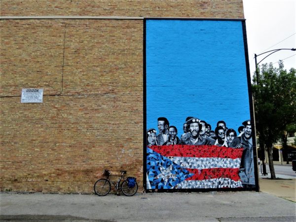 A tour bicycle leaning next to a mural with a blue background and yelling young people standing behind a Puerto Rican flag.