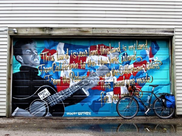 A tour bike leaning on a a garage painted with a portrait of Woody Guthrie and the lyrics to This Land is your Land.