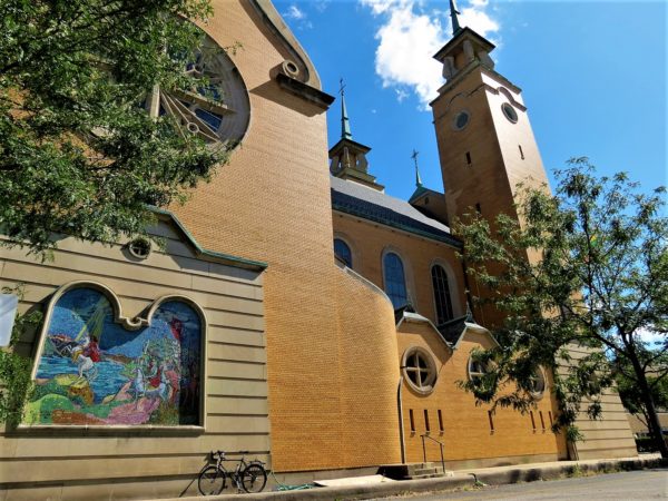 A tour bicycle leaning on the side of a yellow brick church with two front towers and a mosaic on the side