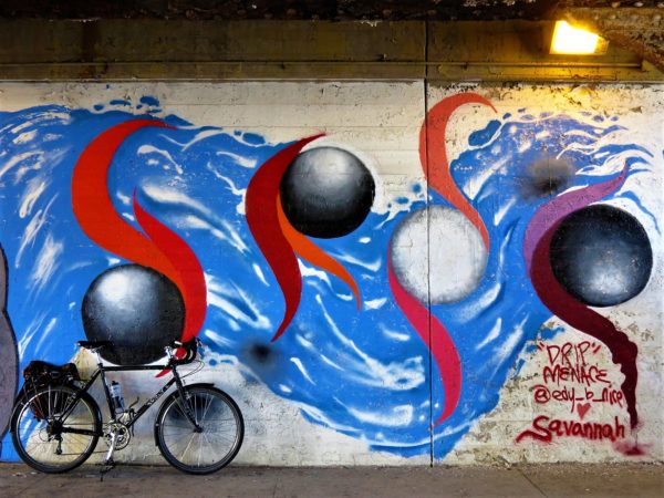 A tour bike leaning on a mural with blakc and white smooth ball with red tail like bits on a blue river like background