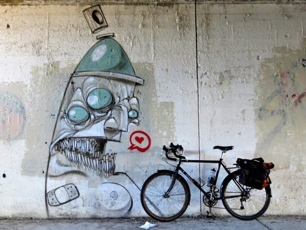 A tour bike leaning next to a grey and pale green mural of an angry sprat paint can with a red heart in a red speech bubble